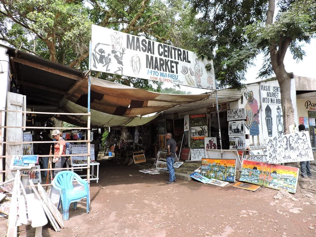 Paintings and carvings on display beneath a canopy at a craft market whose sign reads “Masai Central Market, Mto wa Mbu”